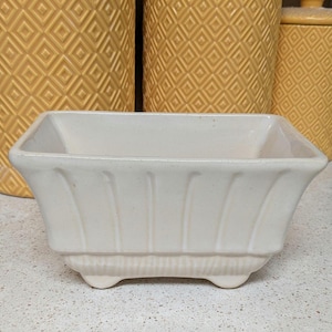 May include: A rectangular, off-white ceramic planter with a fluted design on the sides and a ribbed base. The planter sits on three small, rounded feet. In the background are yellow ceramic canisters.
