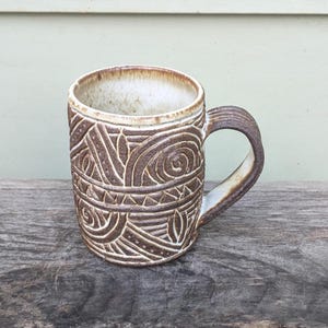 May include: A white ceramic mug with a brown, abstract design. The mug has a rounded handle.