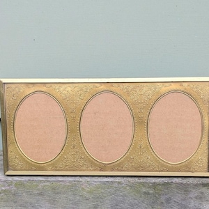 May include: A rectangular, gold-toned picture frame with three oval openings. The frame has an ornate, embossed floral design around the edges. The background is a light blue-green color.