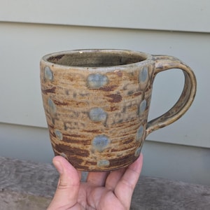 May include: A ceramic mug with a textured, earthy brown and tan surface featuring horizontal ridges and scattered light blue circular spots. The mug has a wide, curved handle and a thick rim, showcasing a rustic, hand-thrown appearance.