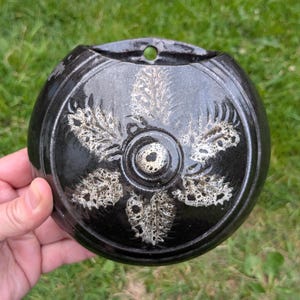 May include: Black ceramic wall pocket with a circular shape and a decorative floral design in white. The wall pocket has a hole at the top for hanging. The object is held in a hand against a background of green grass.