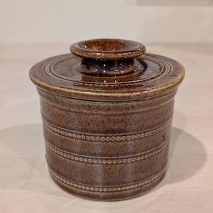 May include: A brown ceramic butter dish with a lid. The dish is cylindrical with horizontal bands of dotted and patterned detailing. The lid has a central knob and a decorative ring around the edge.