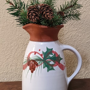 May include: White ceramic pitcher with a brown rim and handle, decorated with a Christmas design of holly, pine needles, and red ribbon. The pitcher holds pine branches and pine cones, creating a festive holiday arrangement.