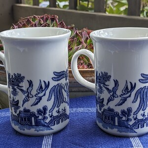 May include: Two white ceramic mugs with blue and white willow pattern. The mugs have a handle and are sitting on a blue and white checkered surface.