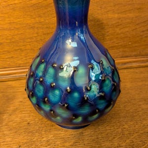 May include: A blue ceramic vase with a bulbous body and a narrow neck. The vase has a textured surface with teal and dark blue accents and small, dark, raised dots. The vase is displayed on a wooden surface.