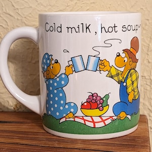 May include: White ceramic mug featuring a cartoon illustration of two bears toasting with mugs, with the text "Cold milk, hot soup." One bear wears a blue polka-dot nightgown and the other wears overalls. A picnic basket with fruit is also shown.