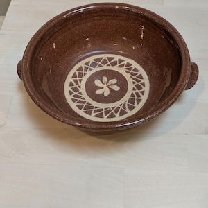 May include: A brown ceramic bowl with a cream-colored floral design in the center. The bowl has two small handles and is set on a light-colored wooden surface. This decorative bowl is suitable for serving.