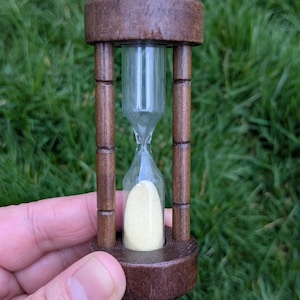 May include: A small hourglass with a dark brown wooden frame and base. The clear glass chamber holds yellow sand. The hourglass is held against a backdrop of green grass.