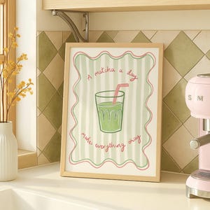 May include: A framed print with the text "A matcha a day makes everything okay." The artwork features a green matcha drink in a glass with a pink straw, set against a striped background. The frame is light brown.