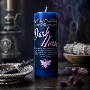 May include: A cylindrical blue pillar candle featuring a black label with the text Kentucky Conjure Blessing Collection Dark Horse. The label includes a pink moth illustration and printed text about blessed wind, steed, and success.