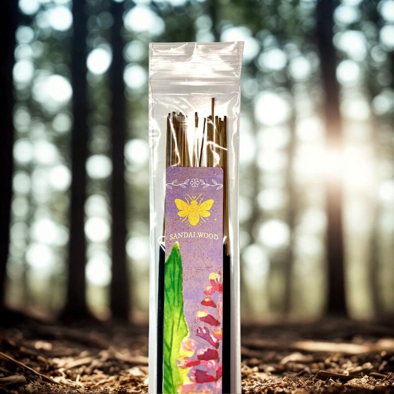 May include: A transparent packet of sandalwood incense sticks. The label displays a yellow bee design and the word "Sandalwood". The background is a blurred forest scene with sunlight.
