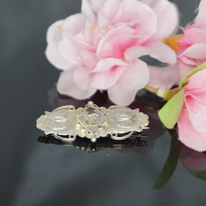 May include: A silver brooch with an intricate design featuring a central diamond-shaped element and two smaller, oval-shaped elements on either side. The brooch is set against a black background with pink flowers.