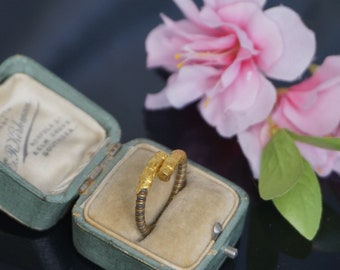 Puede incluir: Un anillo de oro con una superficie texturizada se encuentra dentro de una caja de anillo verde. La caja está abierta y el anillo es visible. Hay flores rosas con hojas verdes en el fondo.