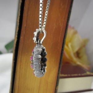May include: A silver pendant necklace with a textured, circular design. The pendant features small, round, pink gemstones and is suspended from a delicate chain. The necklace is displayed against a backdrop of a book and a soft, blurred background.