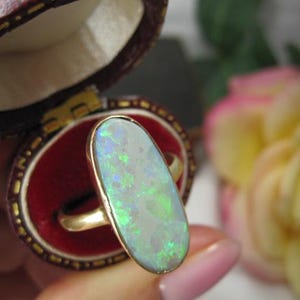 May include: A gold ring featuring a large, oval-shaped opal gemstone with iridescent green and blue flashes. The ring is set in a simple gold band and is positioned within a small, oval-shaped, dark red velvet-lined jewelry box.
