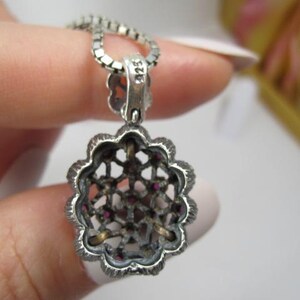 May include: A silver pendant necklace with a floral design. The pendant has a filigree pattern with small red gemstones. The chain is a delicate silver link. The pendant is held by a hand with a white nail polish.