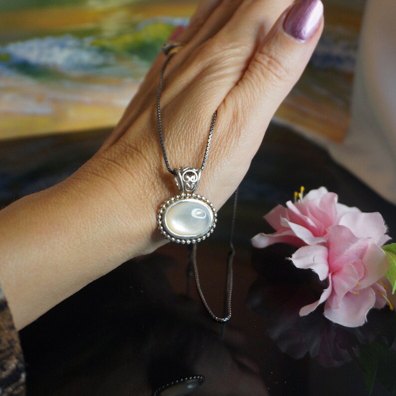 May include: A silver chain necklace with a white oval pendant. The pendant has a beaded border and a silver bail.