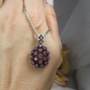 May include: A silver pendant necklace featuring a cluster of small, round, deep pink gemstones. The pendant is a floral-like design with a silver chain. The necklace is held in a hand.