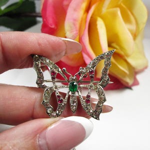 Peut inclure: Broche papillon argentée ornée de pierres précieuses claires et rouges, avec une pierre verte centrale. La broche est tenue dans une main, avec une rose en arrière-plan.