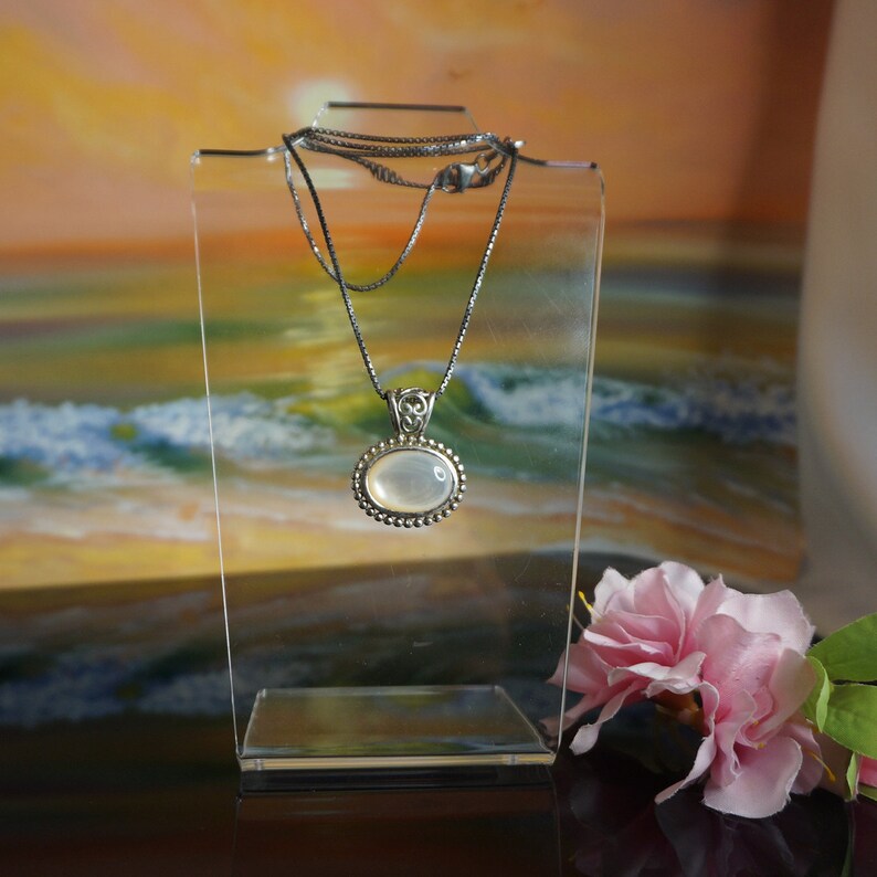May include: A silver chain necklace with a white oval pendant. The pendant is set in a silver frame with a decorative design. The necklace is displayed on a clear stand against a backdrop of a sunset over the ocean.