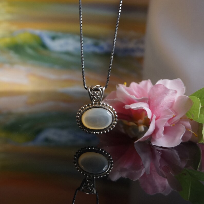 May include: A silver chain necklace with a white oval pendant set in a silver filigree frame. The pendant is surrounded by a row of small silver beads.