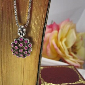 May include: A silver pendant necklace with a floral design, featuring small, round, pink gemstones. The necklace hangs against a backdrop of a vintage book and a blurred yellow and pink flower.