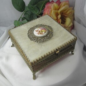 May include: A square jewellery box featuring a cream-coloured, textured lid with a central circular porcelain medallion depicting a floral scene. The box has an ornate, gold-toned metal mesh body with decorative filigree edges and four small feet.
