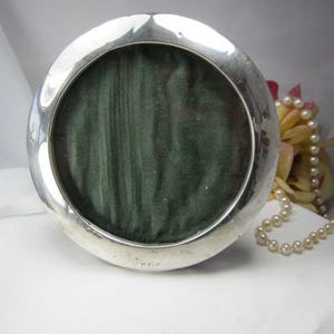 May include: A circular silver picture frame with a polished metallic finish and a smooth, rounded border. The center of the frame is filled with a dark green, textured fabric backing. Small, faint hallmarks are visible at the bottom edge.