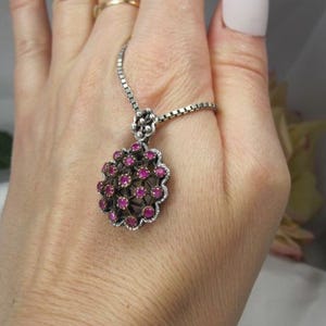 May include: Silver-toned pendant necklace featuring a cluster of small, round, ruby-colored gemstones. The pendant has a floral design with a delicate chain. The necklace is displayed on a hand.