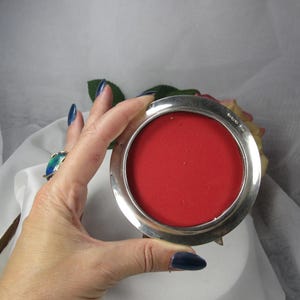 May include: A circular silver-toned metal coaster or small tray featuring a vibrant red felt insert. The metallic rim has a polished, reflective finish and includes small, stamped hallmark engravings along the upper right edge of the frame.