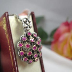May include: A silver pendant necklace with a cluster of small, round, pink gemstones. The pendant is set in a silver and gold-toned metal. The necklace is displayed on a small, red book.