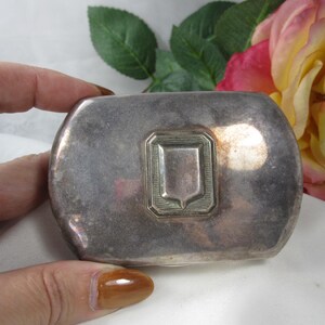 May include: A rectangular silver-toned metal case with rounded corners and a polished finish. The centre of the lid features a raised, shield-shaped emblem surrounded by a rectangular border with fine, etched line detailing on the surface.