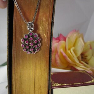 May include: A silver necklace with a pendant featuring a cluster of small, round, pink gemstones. The pendant is suspended from a delicate chain. The necklace is displayed against a backdrop of a vintage book and a yellow and pink rose.