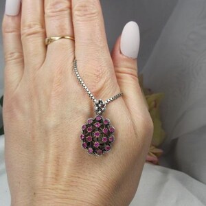 May include: A silver necklace with a round pendant. The pendant is adorned with small, round, pink gemstones set in a floral pattern. The necklace is held in a hand with a gold ring.