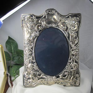 May include: A silver-toned metal picture frame with an oval center opening. The frame features an ornate, raised relief design depicting cherubs, floral motifs, and scrolling patterns throughout the border. The central area is dark blue.