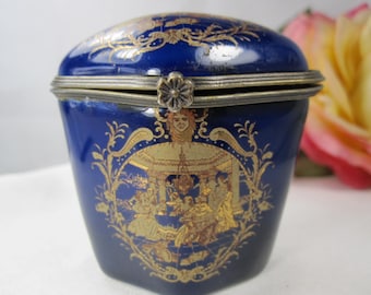 May include: A deep blue porcelain trinket box with a hinged lid and a metallic flower-shaped clasp. The exterior features intricate gold-toned illustrations of figures in a garden setting, framed by ornate scrollwork and a central face motif.
