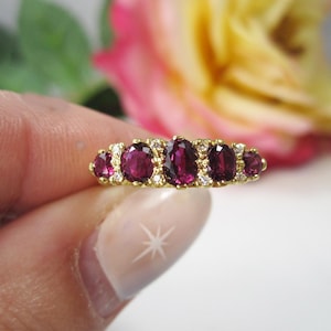 May include: A gold ring featuring a series of oval-cut, deep red gemstones, interspersed with smaller, clear gemstones. The ring's band is a polished gold, and the gemstones are set closely together. The background includes a blurred rose.