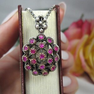 May include: A silver pendant necklace with a floral design, featuring small, round, bright pink gemstones. The pendant is displayed on a light-colored surface, with a blurred background of a pink and yellow flower.