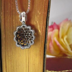 May include: A silver pendant necklace with a floral design. The pendant has a filigree pattern with small red accents. The necklace is displayed on a book with gold-colored pages, with a blurred yellow and pink flower in the background.
