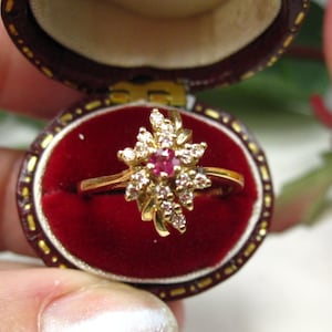 May include: A gold ring with a starburst design, featuring a central red gemstone surrounded by smaller clear stones. The ring is displayed in a burgundy velvet-lined jewellery box.