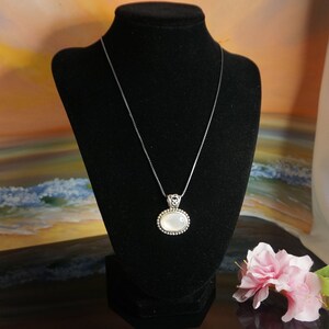 May include: A silver chain necklace with a white oval pendant. The pendant has a silver border with small beads around the edge.