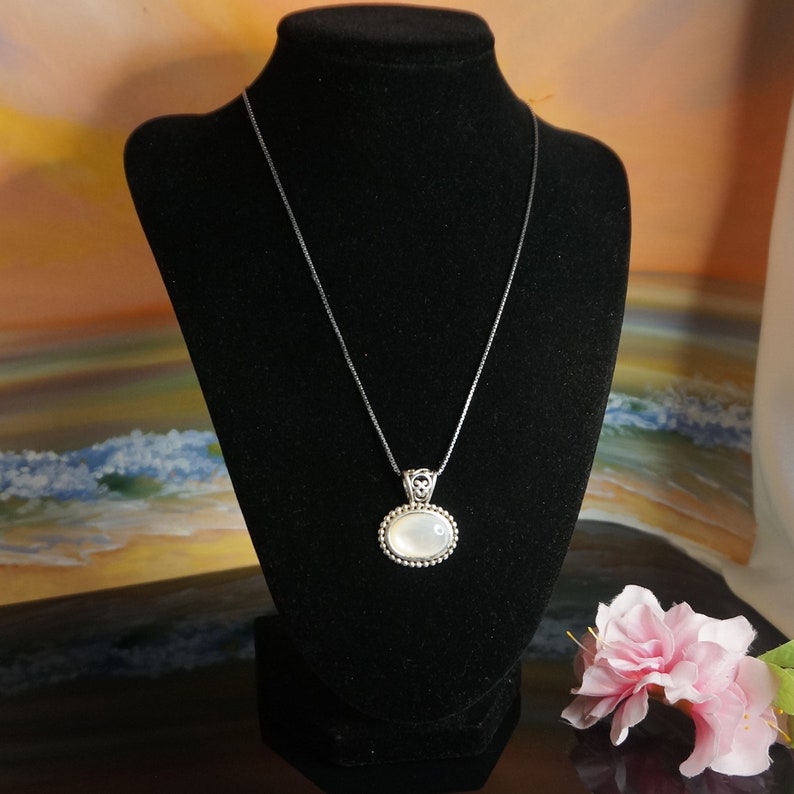 May include: A silver chain necklace with a white oval pendant. The pendant has a silver border with small beads around the edge.