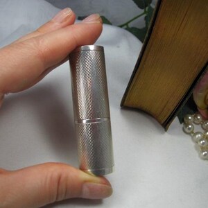 May include: A cylindrical silver-toned metal case featuring a fine cross-hatched, textured pattern across its entire surface. The object is divided into two sections by a horizontal line, indicating a removable cap and a base component.