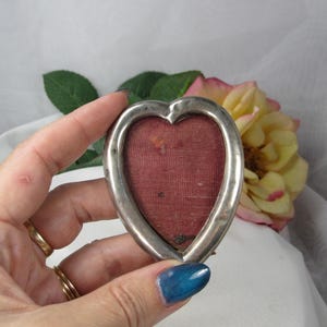 May include: A heart-shaped picture frame with a polished silver-toned metal border and a dark red fabric backing. The frame features a smooth, rounded edge and a central opening designed to hold a small photograph or keepsake item.