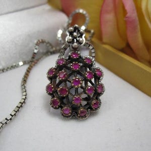 May include: A silver pendant necklace with a detailed, openwork design. The pendant features a cluster of small, round, ruby-colored gemstones. The chain is a delicate, square-link style. The necklace is displayed on a white surface.