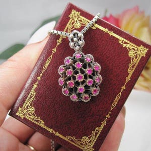 May include: A silver pendant necklace with a floral design, set with small, round, pink gemstones. The pendant hangs on a delicate chain and rests on a small, red book with gold decorative trim.