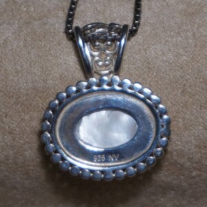 May include: Silver pendant necklace with a round, white, oval-shaped stone set in a silver bezel. The pendant has a decorative border of small silver beads and the back is engraved with "925 NV".
