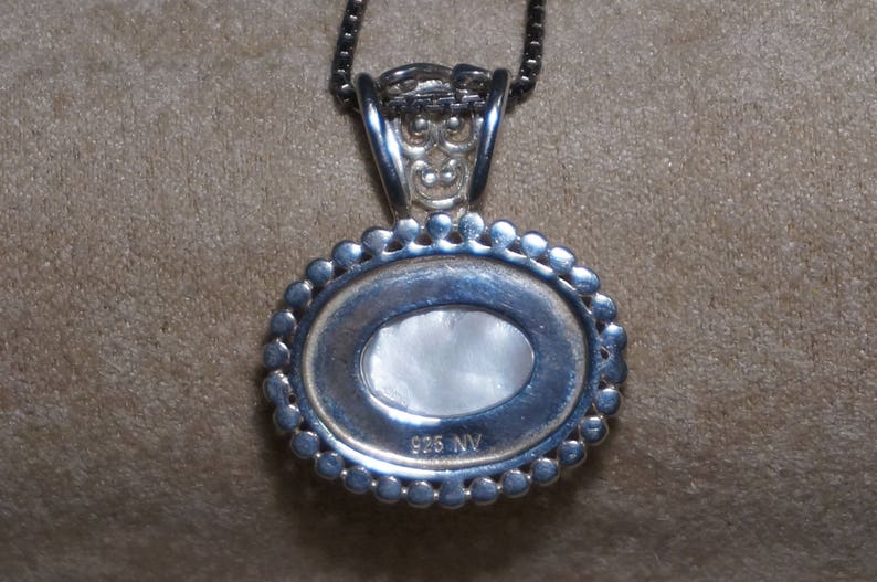 May include: Silver pendant necklace with a round, white, oval-shaped stone set in a silver bezel. The pendant has a decorative border of small silver beads and the back is engraved with "925 NV".
