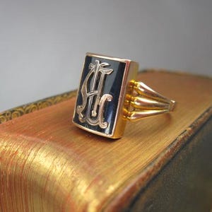 May include: A gold ring featuring a rectangular face with a black enamel inlay. The center of the face displays an ornate, raised gold monogram with the letters AH. The ring band consists of three distinct gold prongs on each side of the face.
