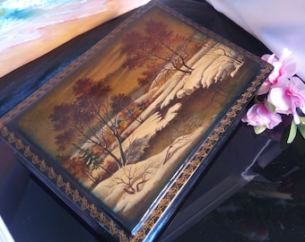 May include: A black wooden box with a gold trim and a painted scene of a winter landscape. The scene depicts a snowy forest with trees and a river. The box has a gold inscription that reads "Winter 1972".